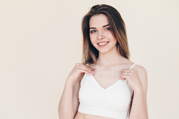 Young Woman in White Sports Top adjusts straps of her bodice. She Stands on Beige Background
