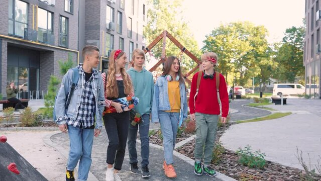 Group of joyful teens friends speaking while walking in town holding in hands skateboards. Stylish Caucasian boys and girls skaters going to skate park. Skateboarding, youth culture, leisure concept