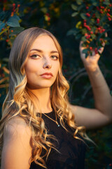 Young beautiful woman in a black dress holds a branch with berries in her hand and looks at the camera. Outdoor portrait in the evening park