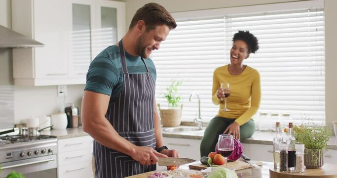 Video of happy diverse couple preparing meal, drinking wine and having fun in kitchen