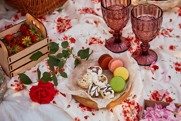 Summer aesthetic pink picnic with tablecloth marshmallows, macaroons, strawberries, glasses of wine among pink roses in the park. Romantic dinner.