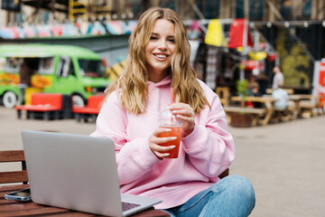 A young woman in a pink hoodie is working at a laptop and drinking lemonade. Against the background of a hipster street food court