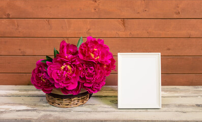 Red peony flowers in wicker basket and empty white frame on wooden table.