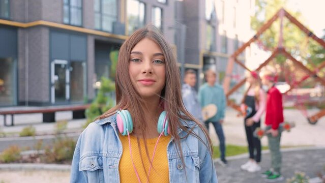 Camera approaching cheerful teen girl with headphones standing outdoors looking at camera and smiling. Happy female teenager on street. Smile, positive emotions, close up concept Friends on background