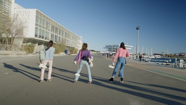 Three girls roller skate on road by water while holding their shoes
