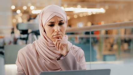 Young pensive puzzled arab woman in hijab working on laptop concentrating on idea looking for...