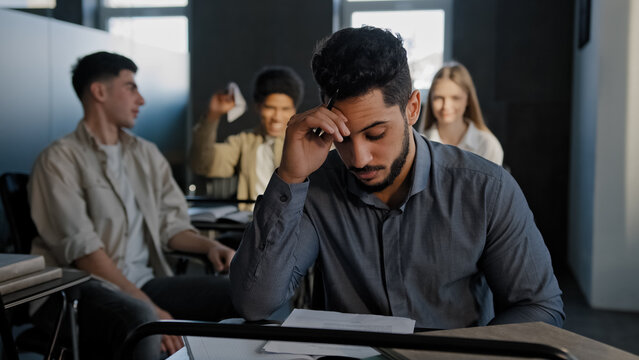 Frustrated oppressed indian student sitting in class lonely young guy suffers from abuse bullying by classmates ridicule mockery bad attitude upset worried man feeling offended disappointed depression