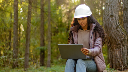 Attractive woman forestry engineer in protective helmet enters data into laptop takes reforestation action young experienced female specialist ecologist technician watching nature reserve checks trees - obrazy, fototapety, plakaty