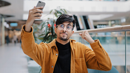 Young happy indian guy blogger holds smartphone recording vlog filming video for blog in mall...