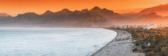 Sunset panoramic view of scenic and popular Konyaalti beach in Antalya resort town. Majestic mountains with haze in the background. Vacation and holiday in Turkey - obrazy, fototapety, plakaty