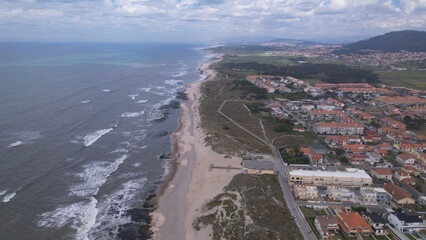Fototapeta premium Aerial view of the Northern Littoral Natural Park in Esposende, Portugal. Sea, beach boulders, pebble shore, and waves. Sea waves breaking on rocks. Sunny beach with sand dunes and a cloudy sky.