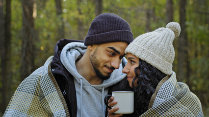 Young happy married couple on date in nature guy and girl lovers in autumn forest embracing in warm blanket drinking tea communicate talk cute hugging enjoy time spent together warm up with hot drink