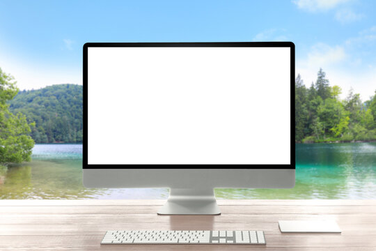 Modern computer with blank screen on wooden table and view of beautiful river and mountains