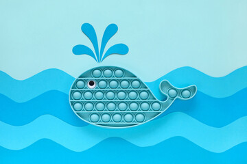 Blue whale releases a fountain of water in blue sea waves made of color paper cut out. Pop it toy, popular antistress sensory silicone bubble fidget toy. Ocean vacations. Summer sea holidays concept.