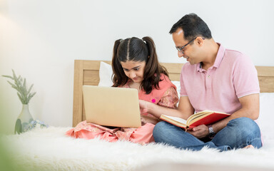 Fototapeta premium Indian happy father and daughter sitting on bed at cozy home, smiling with happiness, teaching, doing homework, playing, using laptop together. Education, Technology, Creative Activity Concept.