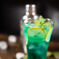a bright green cocktail with lemon and mint in a large transparent glass, mint and ice are scattered on a wooden table with free space for you text