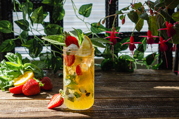 citrus cold lemonade with ice in a beautiful glass on the background of the window, decorated with strawberries, a slice of lemon and mint, sprigs of mint and strawberries in the background, summer