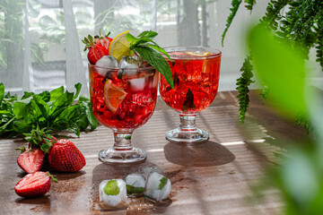 two glasses of strawberry cold lemonade with ice on the window background, decorated with strawberries, lemon slice and mint, sprigs of mint and strawberries in the background, summer heat, delicious