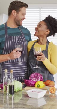 Vertical video of happy diverse couple preparing meal in kitchen and drinking wine