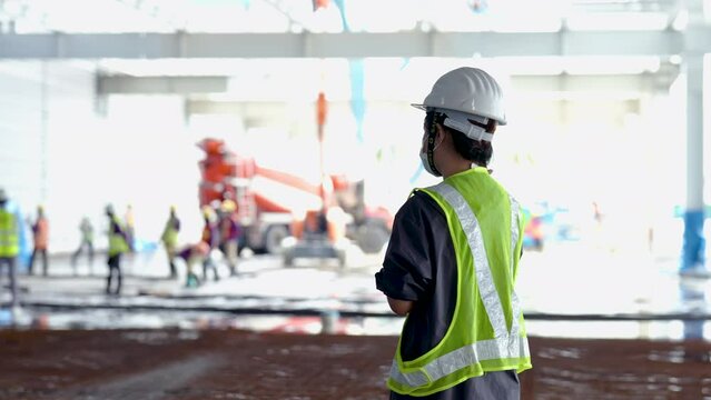 4K, An Asian female engineer wearing a reflective green safety vest wears a protective hard hat. Standing supervisor at a construction site with construction workers working in the background.