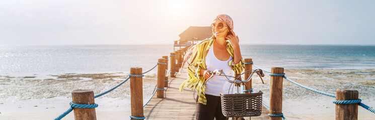 Portrait of a smiling woman dressed in light summer clothes, sunglasses with bicycle on the wooden sea pier on the sandy Zanzibar beach.Careless vacation in the tropical countries concept image