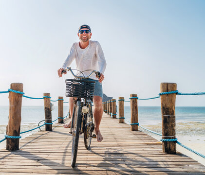 Portrait of a happy smiling man dressed in light summer clothes and sunglasses riding a bicycle on the wooden sea pier. Careless vacation in tropical countries concept image.