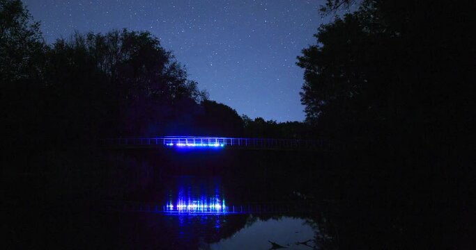 The movement of stars in the night sky over the bridge with colored backlighting, timelapse of the night landscape. Silhouette of trees.