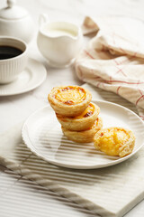 Traditional portuguese vanilla pudding puff pastry pastel de nata stacked on each other on white plate on marble board with a cup of coffee in white porcelain