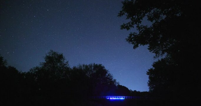 The movement of stars in the night sky over the bridge with colored backlighting, timelapse of the night landscape. Silhouette of trees.
