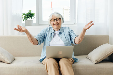 an elderly woman with gray hair is sitting on a sofa with a laptop using headphones, in a comfortable environment and actively gesticulating with her hands spreading them apart looking at the monitor