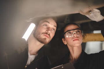 Fototapeta premium Young male and female car mechanic wearing safety glasses and hand gloves standing under car in garage inspecting and doing maintenance work for car