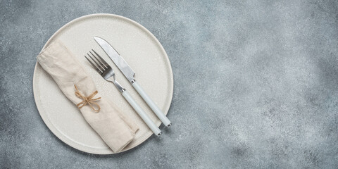 Clean beige plate with cutlery and linen napkin, gray grunge background. Table setting. Top view,...