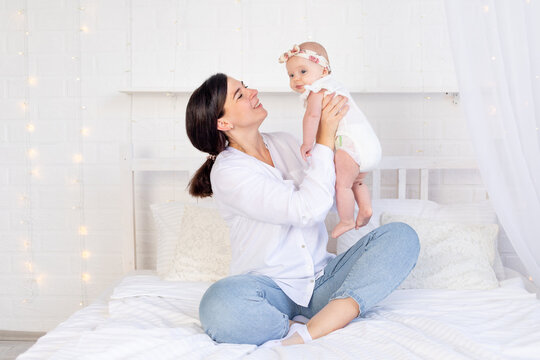 mom and baby girl hug and kiss sitting on a white cotton bed at home, maternal love and care