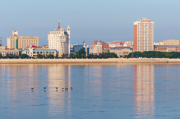 Naklejka premium View of China from Russia across the Amur River on a summer morning. A flock of cormorant birds are flying over calm water. City buildings of different styles and purposes. Clear sky.