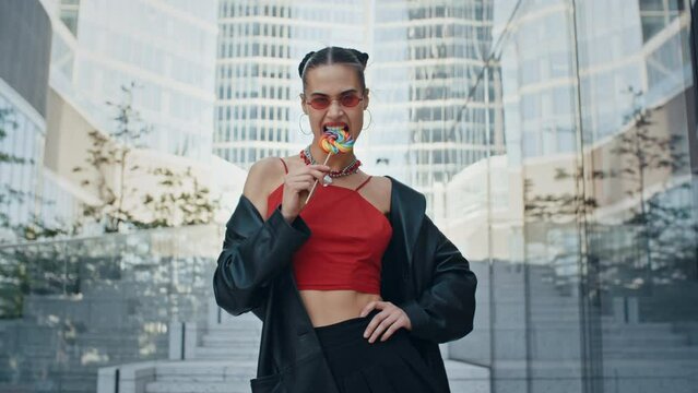 Gorgeous happy woman seductively bites big multi-colored lollipop and smiles, stylish teen model wearing cool red eye glasses posing with colorful candy standing in modern city street