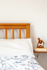 Wooden bed back, with white sheets, bedside table with books and small wooden figure. 