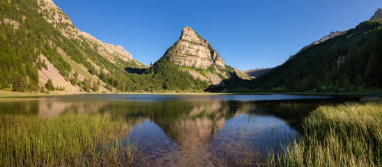 Lac des Sagnes in Summer with La Tour des Sagnes pyramid-shaped mountain in the Mercantour National Park (panoramic) at sunset. Jausiers, Ubaye Valley, Alpes de Haute Provence, Alps, France - 515249895