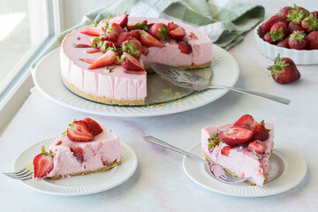 Slices of strawberry cheesecake with the cake in behind, against a bright window