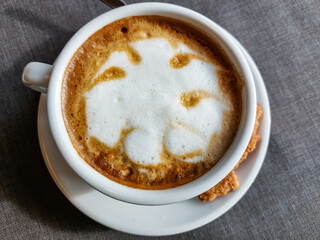 View of a mug of a cappuccino coffee with fluffy, white milk froth on the table covered with neutral grey tablecloth in front of window