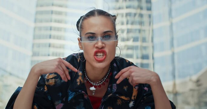 Urban style cheeky female wearing colored windbreaker and cool futuristic glasses rapping aggressively while sitting on stairs in downtown overlooking high-rise skyscrapers. Handsome fashion model