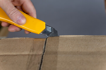 A man's hand opens a cardboard box with a yellow cutter. The flaps of the package are sealed with adhesive tape. Close-up. Selective focus.
