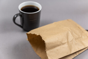 A brown paper bag and a cup of coffee on gray. A full cup of hot, invigorating drink. A lunch bag of food. Selective focus.