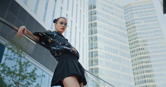 Urban style seductive girl wearing colored jacket short skirt and cool futuristic glasses posing leaning back against glass parapet near high rise skyscrapers. Handsome female fashion model
