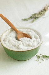 Salt in a ceramic bowl with a wooden spoon and a sprig of dry rosemary on the table. Rustic style. Natural resource. Vertical orientation.