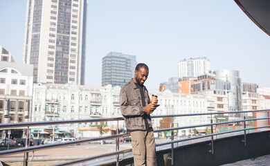 Joyful travel blogger enjoying mobile messaging in social media during leisure for cell networking in city, cheerful African American hipster guy with coffee to go browsing website on smartphone
