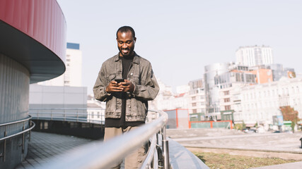 Cheerful dark skinned man in bluetooth earbuds listening positive audio music while chatting with network followers in social media, African American travel blogger enjoying playlist from cellphone