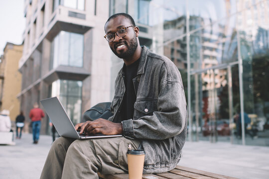 Portrait of skilled digital nomad in classic eyewear smiling at camera during time for working remotely on netbook at city street, happy dark skinned man in spectacles using laptop computer