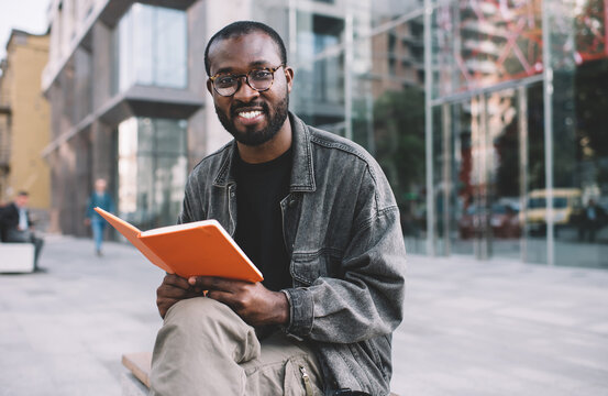 Portrait of carefree male reader in spectacles holding education textbook for learning and posing at city urbanity, cheerful hipster guy in eyewear smiling at camera during leisure with book