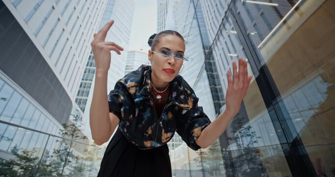 POV wide angle urban style cheeky female wearing colored windbreaker and cool futuristic glasses rapping aggressively in downtown overlooking high-rise skyscrapers. Handsome fashion model