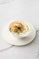 Norwegian red salmon fish soup with vegetables, herbs and cream, bowl of soup on a white marble background